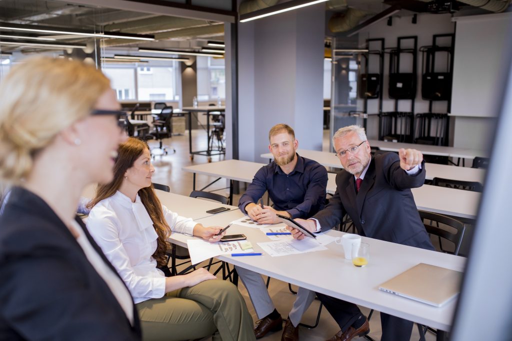 Executivos em reunião estratégica, com um empresário experiente apontando para o flip chart enquanto apresenta ideias para a equipe